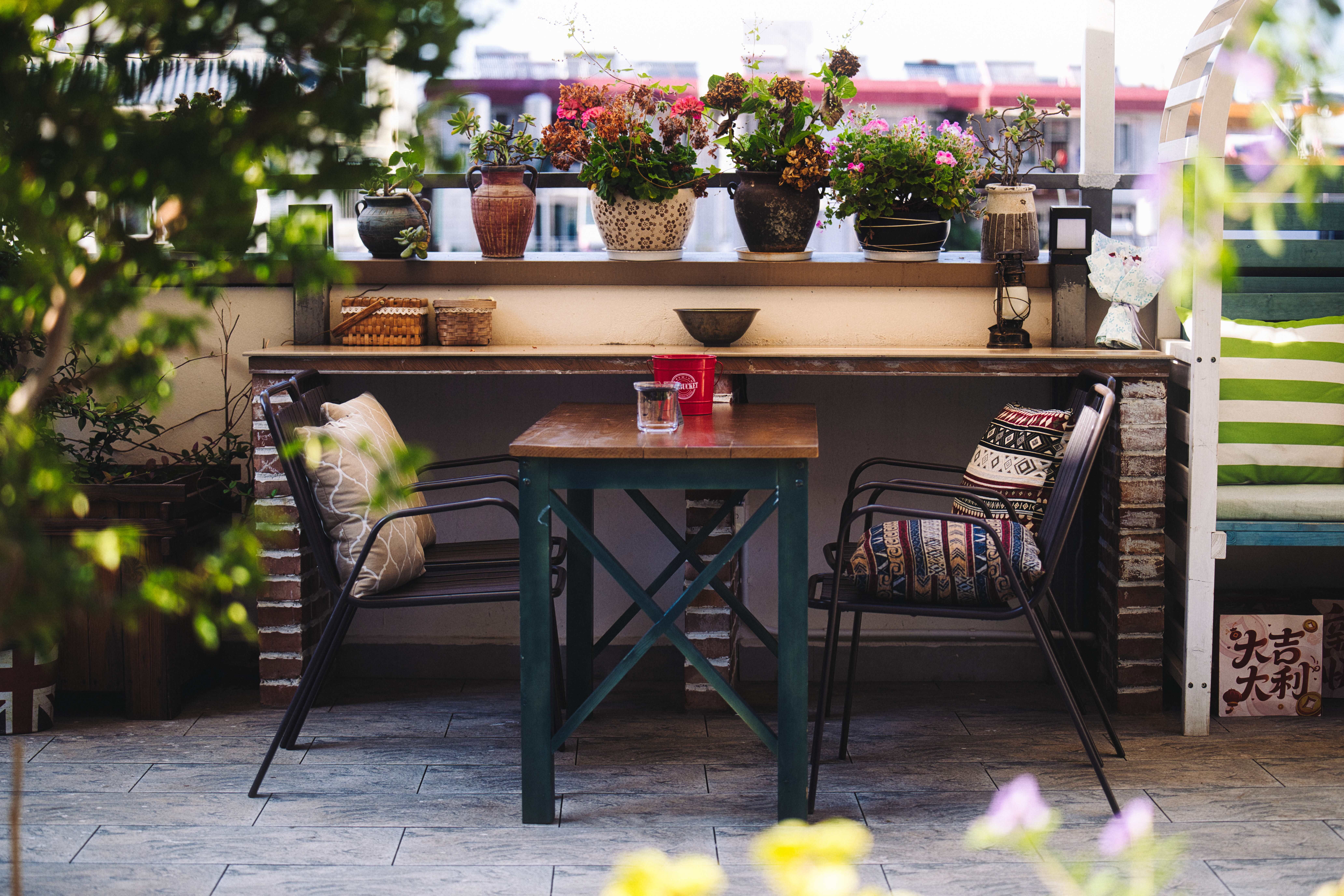 An intimate balcony seating nook with a small wooden table, textured cushions, potted plants, and warm brick walls